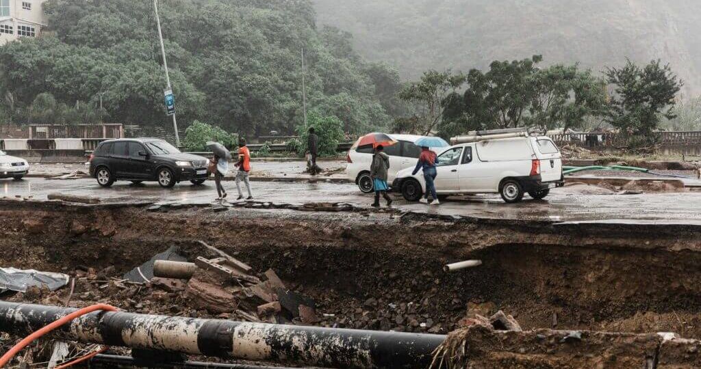 Severe flood damage to a road in South Africa with collapsed infrastructure, exposed pipes, stranded vehicles, and pedestrians during heavy rainfall, illustrating the growing impact of extreme weather on property and infrastructure.