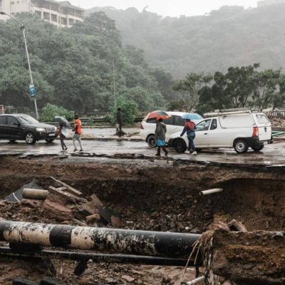 Severe flood damage to a road in South Africa with collapsed infrastructure, exposed pipes, stranded vehicles, and pedestrians during heavy rainfall, illustrating the growing impact of extreme weather on property and infrastructure.